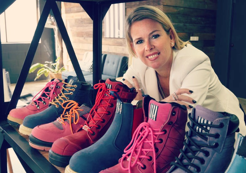 Stacey Head smiling with blonde hair and cream jacket, arms folder with various pink and purple boots on display in front of her