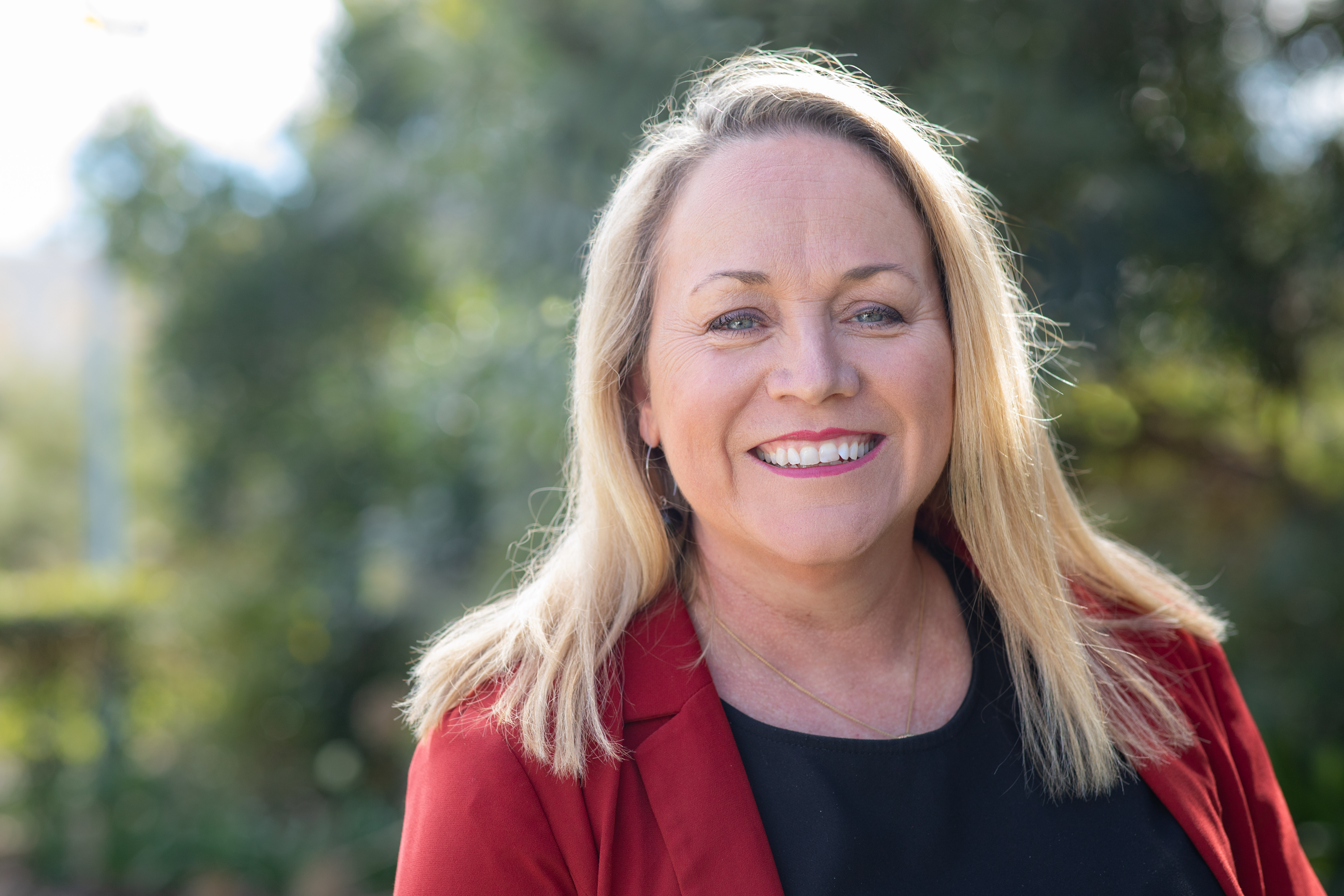 Trudi Bartlett smiling with blonde shoulder length hair, wearing a black shirt and a red jacket in front of a green garden in the background